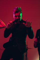 Young and joyful caucasian singer performing on red background in neon light. Concept of music, hobby, festival. Colorful portrait of modern artist. Attented, inspired. Art, cover band. Close up.