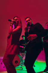 Young and joyful caucasian singers performing on red studio background in neon light. Concept of music, hobby, festival. Colorful portrait of modern artist. Attented and inspired. Art, cover band.