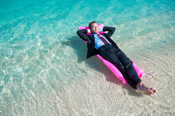 Relaxed businessman floating on a bright pink pool raft in shallow tropical blue waters