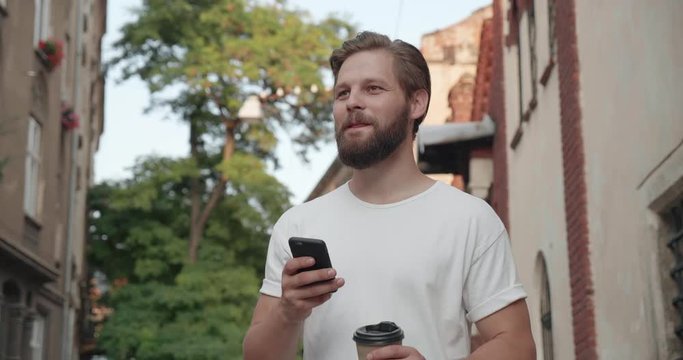 Stylish Young Caucasian Male Using social networks on his Modern Mobile Phone. Having fun while walking in the City. Looking Happy and gorgeous. Drinking Coffee. Man with nice Hairstyle.