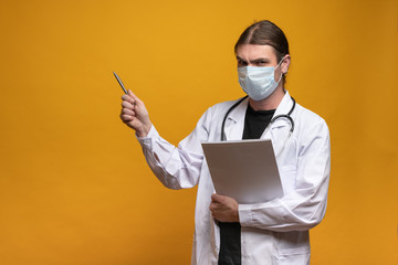 Young doctor holding a pen and a map is displayed while looking angry using a protection mask and stethoscope to show away to the left