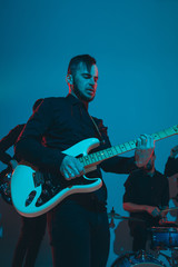 Obraz premium Young caucasian musicians, band performing on blue studio background in neon light. Concept of music, hobby, festival. Colorful portrait of modern artist. Attented and inspired. Art, cover band.