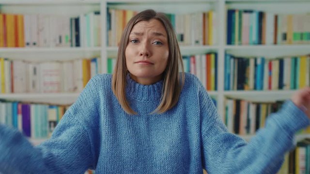 Academic pretty student woman shrugging her shoulders in hesitation shows helpless face cant help or dont understand. posing near bookshelves at library. Doubt, misunderstanding.