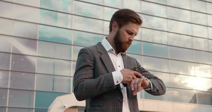 Serious and Busy Businessman Standing Outside Office and Working on his Mobile Device. Bearded Businessman using Smartwatch. Sucessfull People. Technology. Modern Lifestyle.