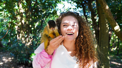 Beautiful curly-haired girl in pink laughs, smiles at saimiri monkey. funny little macaque eats feed from her hands on background of green tropics, rays of sun. Emotional model hold monkey in hands © Artem