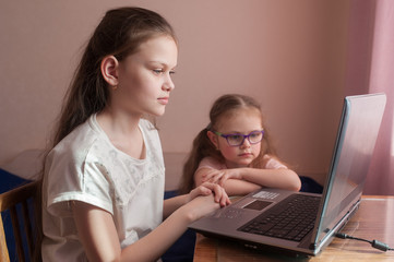 Two girls, the oldest and the youngest, are engaged at a table on a laptop. Little sister with...