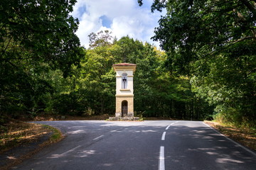 Fototapeta na wymiar Solitary chapel on crossroad in middle of beautiful woods, road traffic, loneliness or coronavirus quarantine concept, sunny day