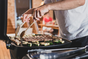 Hands of a man seen flipping cevapcici meal on a grill at an evening party setting. Other guests...