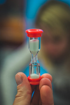 Sand clock in a hand of a man. Hour glass with glass body and red end caps is standing up. Silhouette of a blonde woman in the background.