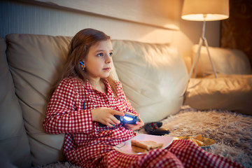 Happy little girl in pajams sitting on sofa, eating pizza and playing video games with joystick at home.