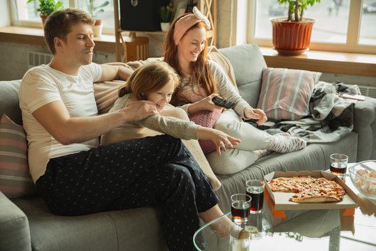 Family Spending Nice Time Together At Home, Looks Happy And Cheerful. Mom, Dad And Daughter Having Fun, Eating Pizza, Watching Sport Match Or TV. Togetherness, Home Comfort, Love, Relations Concept.