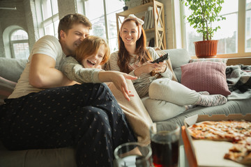 Family spending nice time together at home, looks happy and cheerful. Mom, dad and daughter having fun, eating pizza, watching sport match or TV. Togetherness, home comfort, love, relations concept.