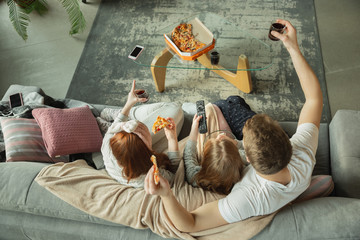 Family spending nice time together at home, looks happy and cheerful. Mom, dad and daughter having fun, eating pizza, watching sport match or TV. Togetherness, home comfort, love, relations concept. - obrazy, fototapety, plakaty