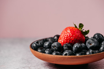 rustic handmade plate full of blueberries with a strawberry on top ready to eat placed on a cement textured background and pink background