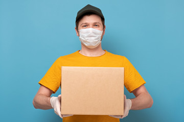 Young delivery man in nedical mask holding and carrying a cardbox on blue wall