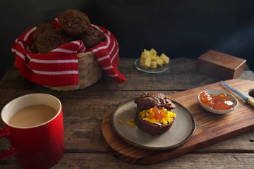 Bran muffin with cheese and apricot jam on a stoneware plate.  Butter knife sits on the edge of the plate.  A small dish of apricot jam sits to the right of the muffin. A cup of tea sits on the table.