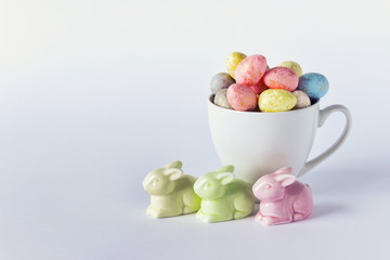 Easter composition greeting card with bunnies rabbits figurines and cup filled with colorful small eggs. Photo with blank space.
