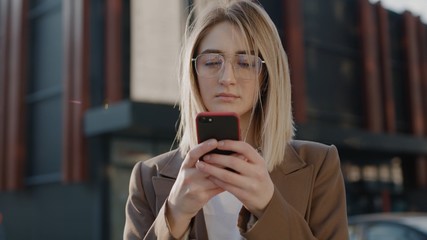 young serious businesswoman in glasses using modern smart phone while walking at break in the city, professional female employer typing text message on cellphone outside spring