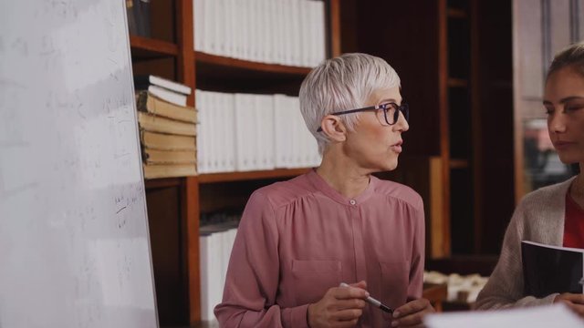 Professor helping student in library at college. Group of multiethnic students taking an active part in a lesson while standing in library. Mature woman teacher and university student discussing.