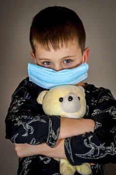 Sad And Tense Cute Caucasian Boy Wearing A Protection Mask Holding A Teddy Bear. No School Depression, Self Isolation Concept, COVID 19 Pandemic Traumatic Experience For A Child