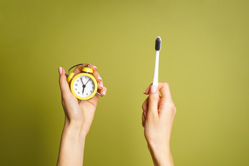 Yellow alarm clock and a toothbrush in the hands of a young woman on a green background. Top view. Morning concept