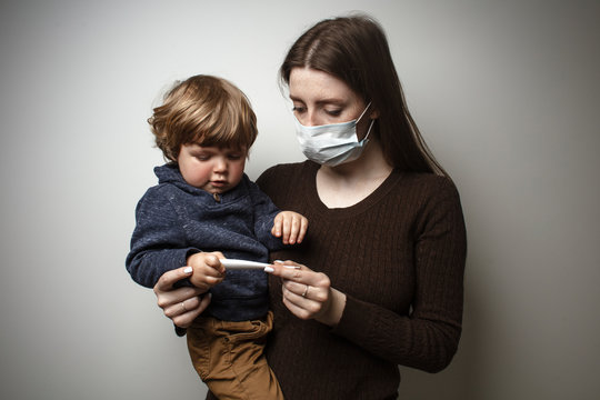 A young woman wearing a face mask, holding in her hands a toddler, and looking at the digital thermometer. Protection against the COVID-19 transmission. Mother and child in quarantine.