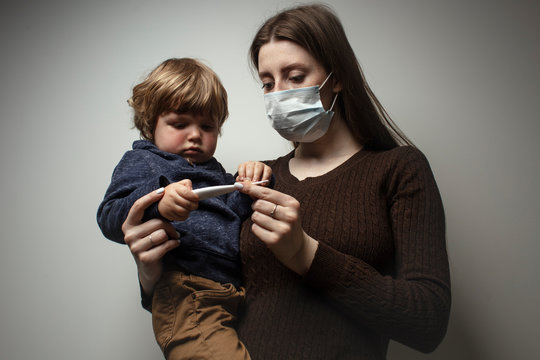 A young woman wearing a face mask, holding in her hands a toddler, and looking at the digital thermometer. Protection against the COVID-19 transmission. Mother and child in quarantine.
