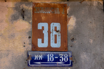 Street number 36 on a metal placard against a stucco wall, white numbers on a red and blue background