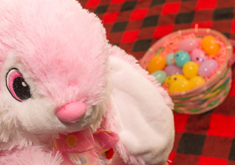 Soft pink easter bunny rabbit sitting on a red plaid blanket with a basket full of colorful eggs