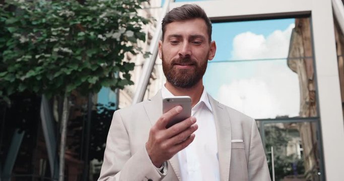 Sucessfull Businessman browsing his Smartphone near modern Office Building. Attractive Man is smiling and wearing elegant Suit Style. Luxury Lifestyle. Successfull Business Man in Crisis.