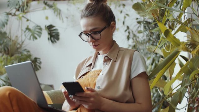 Tilt down shot of young thoughtful woman sitting in armchair, typing on laptop and using smartphone while working in flower shop filled with lots of houseplants