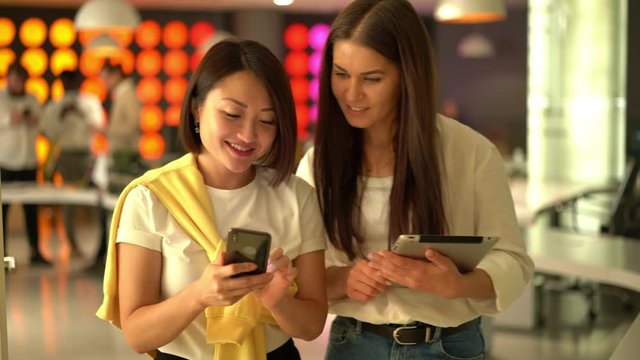 Diverse women in smart casual clothing discussing web publication shared to social networks, millennial colleagues talking about mobility browsing during working day in office interior