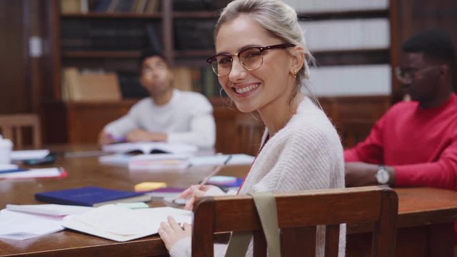 Portrait of university female student studying in library. Happy young woman wearing spectacles studying in college library and looking at camera. Satisfied girl studying for exam.