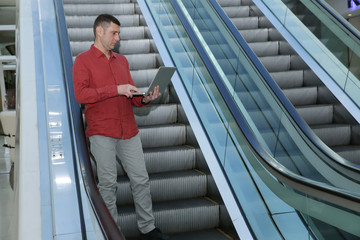 Young handsome businessman in red shirt on an escalator with an open laptop in his hands in a business center of a shopping center or airport.
