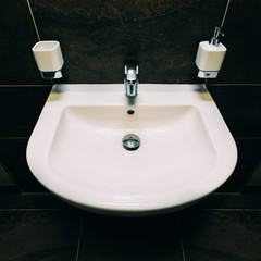 Medium-size U-shaped washbasin in white.Completed with chrome,lever,faucet and plug.There is bathroom accessories like soap dispenser and ceramic cup for brushes.In background are dark colored tiles.