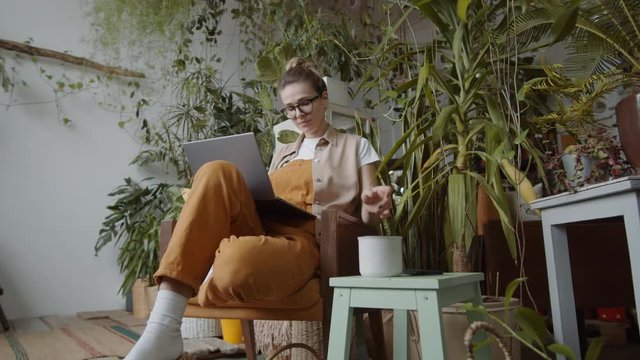 Zoom in shot of young woman sitting in armchair, surfing the Internet on laptop and drinking tea from mug while working in home garden filled with lots of plants