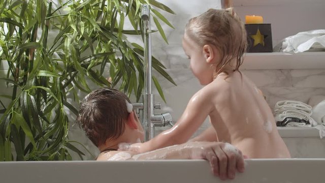 Two children, brother and sister, are bathing in foamy water. The boy is sitting in the bath under the faucet and his younger sister is washing his head. They are both covered in foam. There is a big