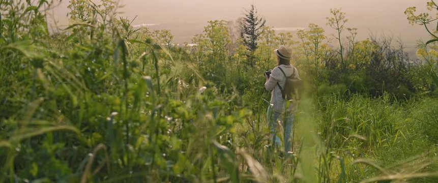 A young girl with backpack and a hat walking through the green field, taking pictures of landscape and nature at sunset. SLOW MOTION, SHALLOW DOF. Adventure, lifestyle concept. BMPCC 4K 