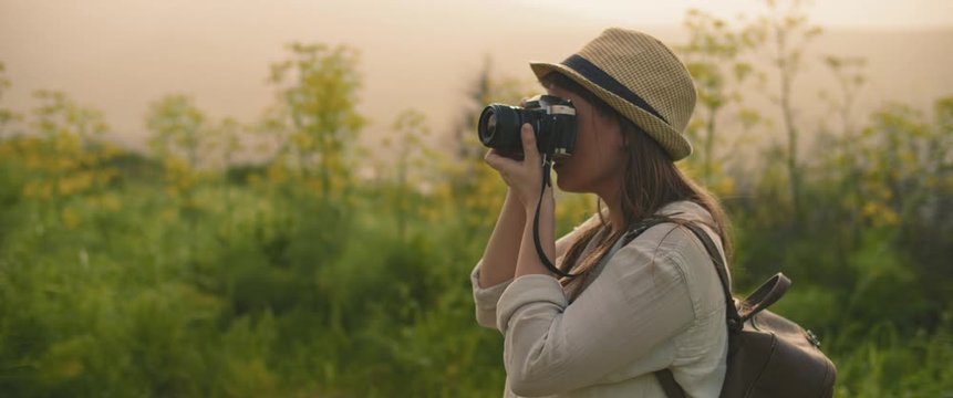 A young girl with backpack and a hat taking pictures of landscape and nature with vintage camera at sunset. CLOSE UP, SLOW MOTION, SHALLOW DOF. Adventure, lifestyle concept. BMPCC 4K 