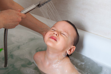 Mom washes her baby's head in a bath with water. Flushing shower shampoo foam. Shampooing without...