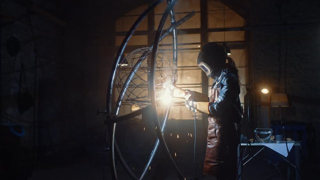 Beautiful Female Artist is Welding a Brutal Metal Sculpture in a Dark Studio. Tomboy Girl Polishes Metal Tube with Sparks Flying Off It. Contemporary Fabricator Creating Abstract Steel Art.