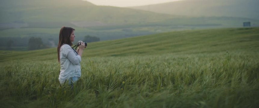 A young girl taking photos of landscape with vintage camera and walking through the wheat field at sunset. SLOW MOTION, SHALLOW DOF. Adventure, lifestyle concept. BMPCC 4K 