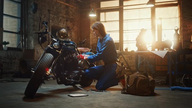 Young Beautiful Female Mechanic is Working on a Custom Bobber Motorcycle. Talented Girl Wearing a Blue Jumpsuit. She Uses a Ratchet Spanner to Tighten Nut Bolts. Creative Authentic Workshop Garage.