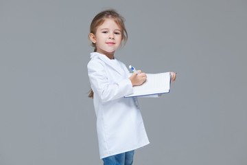 Adorable child girl uniformed as doctor is writing medical record isolated on grey background