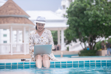 Asian middle age woman using computer for online shopping or social media by the pool at hotel resort in sunny day. Telecommunication device for old people concept