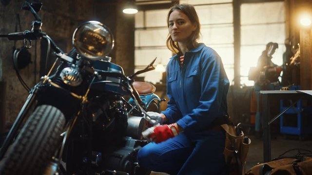 Young Beautiful Female Mechanic is Working on a Custom Bobber Motorcycle. Talented Girl Wearing a Blue Jumpsuit. She Turns and Smiles at the Camera. Creative Authentic Workshop Garage.