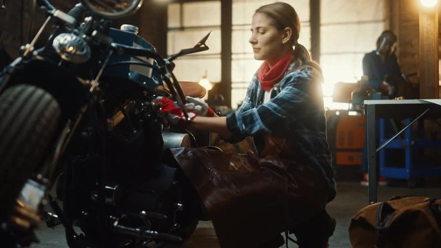 Young Beautiful Female Mechanic is Working on a Custom Bobber Motorcycle. Talented Girl Wearing a Checkered Shirt and an Apron. She Turns and Smiles at the Camera. Creative Authentic Workshop Garage.