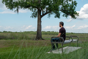 Fototapeta premium A man with a red shirt and blue jeans relaxes on a park bench playing with his phone and admiring the world around him on a bright sunny day on a park bench