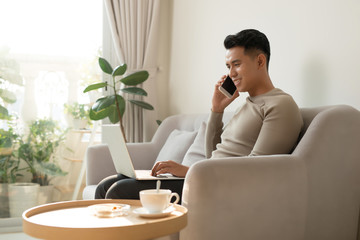 Young man sitting on sofa at home with laptop talking with friend on smartphone