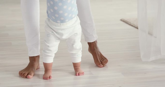 Infant barefoot baby girl boy learning to walk standing on warm floor. African american mum holding hand helping cute little child making first steps at home. Underfloor heating concept, close up view